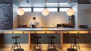 A modern kitchen with a brick backsplash, a chef preparing food, and bar stools. - 9 C Hotel Asahikawa (Asahikawa)