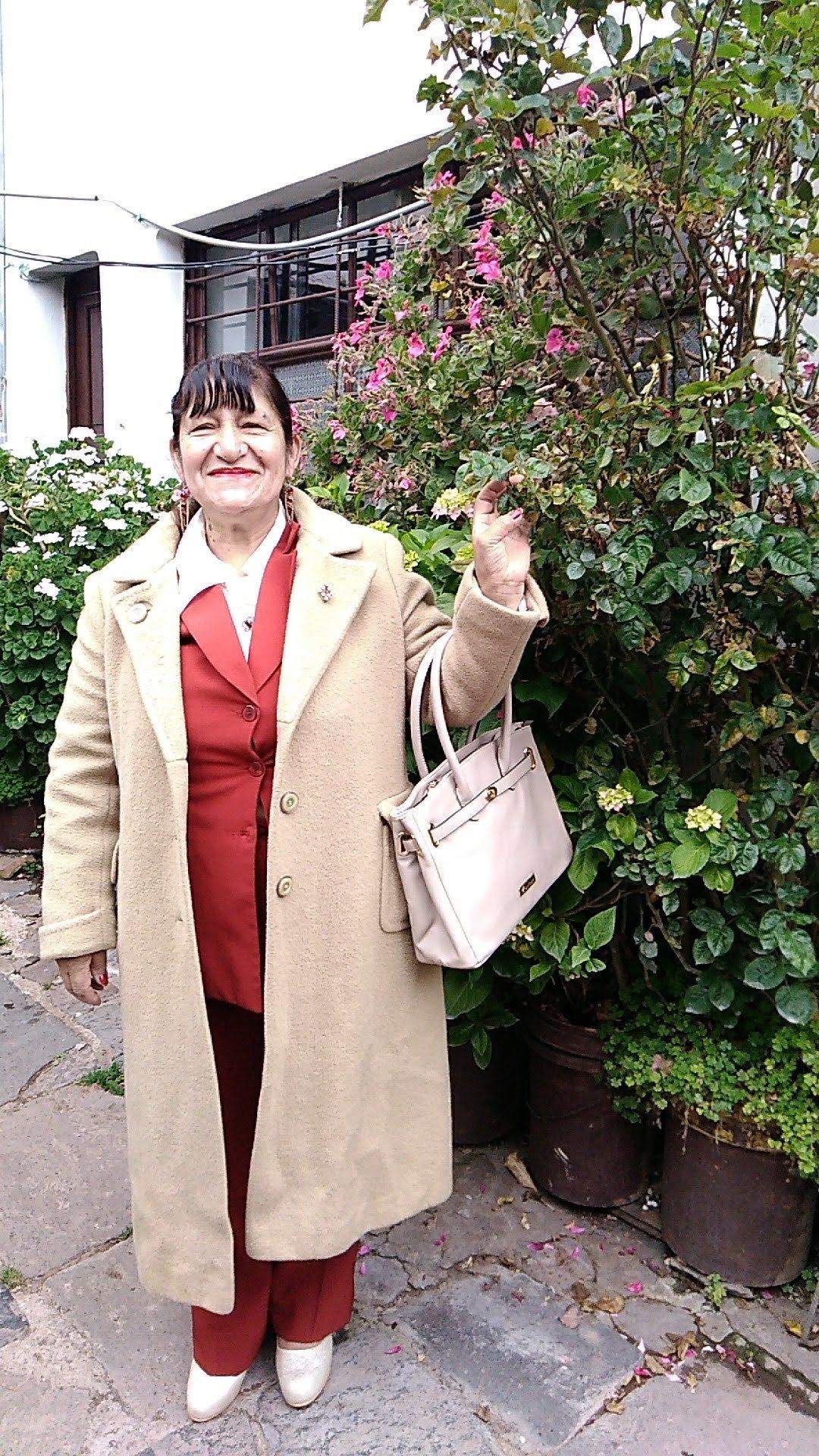 Una persona parada al aire libre, vestida con un abrigo largo color beige, ropa roja y zapatos blancos, sosteniendo un bolso de color claro.