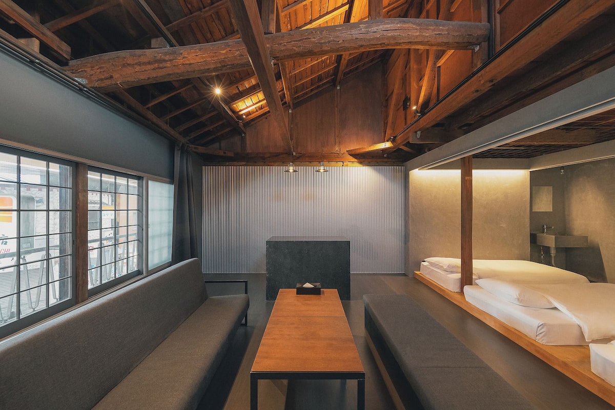 A room with a wooden ceiling, a window with a view of bicycles, a bench, a table, and a bed with white linens.
