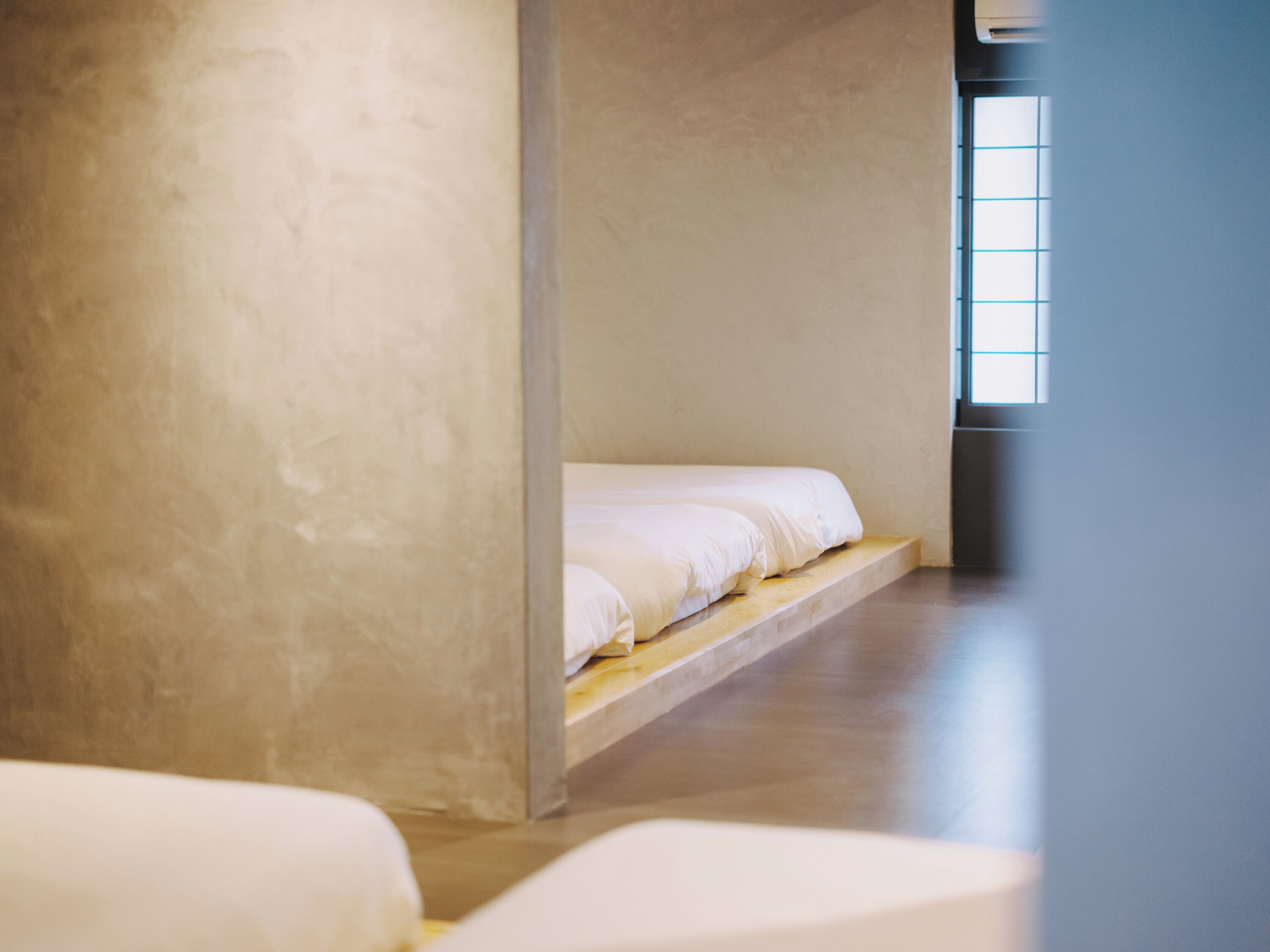 Ein schlicht eingerichteter Raum mit einem Einzelbett, einer Holzplattform und einem großen Fenster.