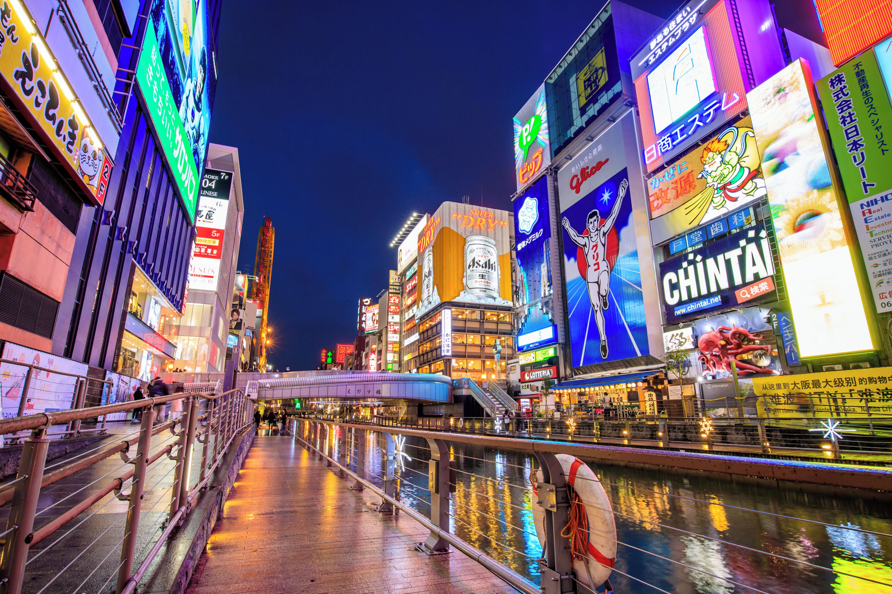 A nighttime cityscape with illuminated billboards and a pedestrian bridge over a canal.