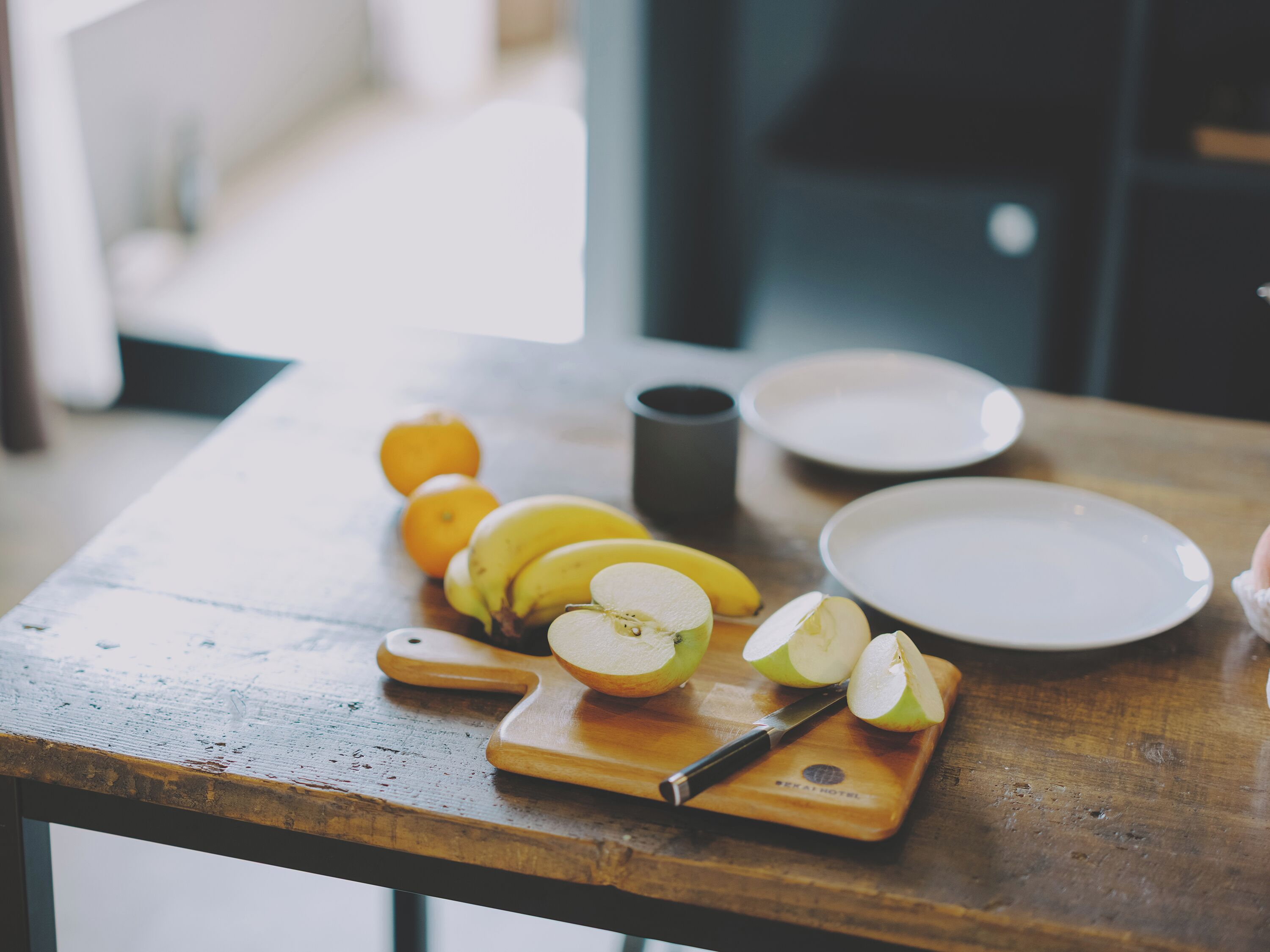 A wooden table with a cutting board, a knife, sliced apples, bananas, and oranges.