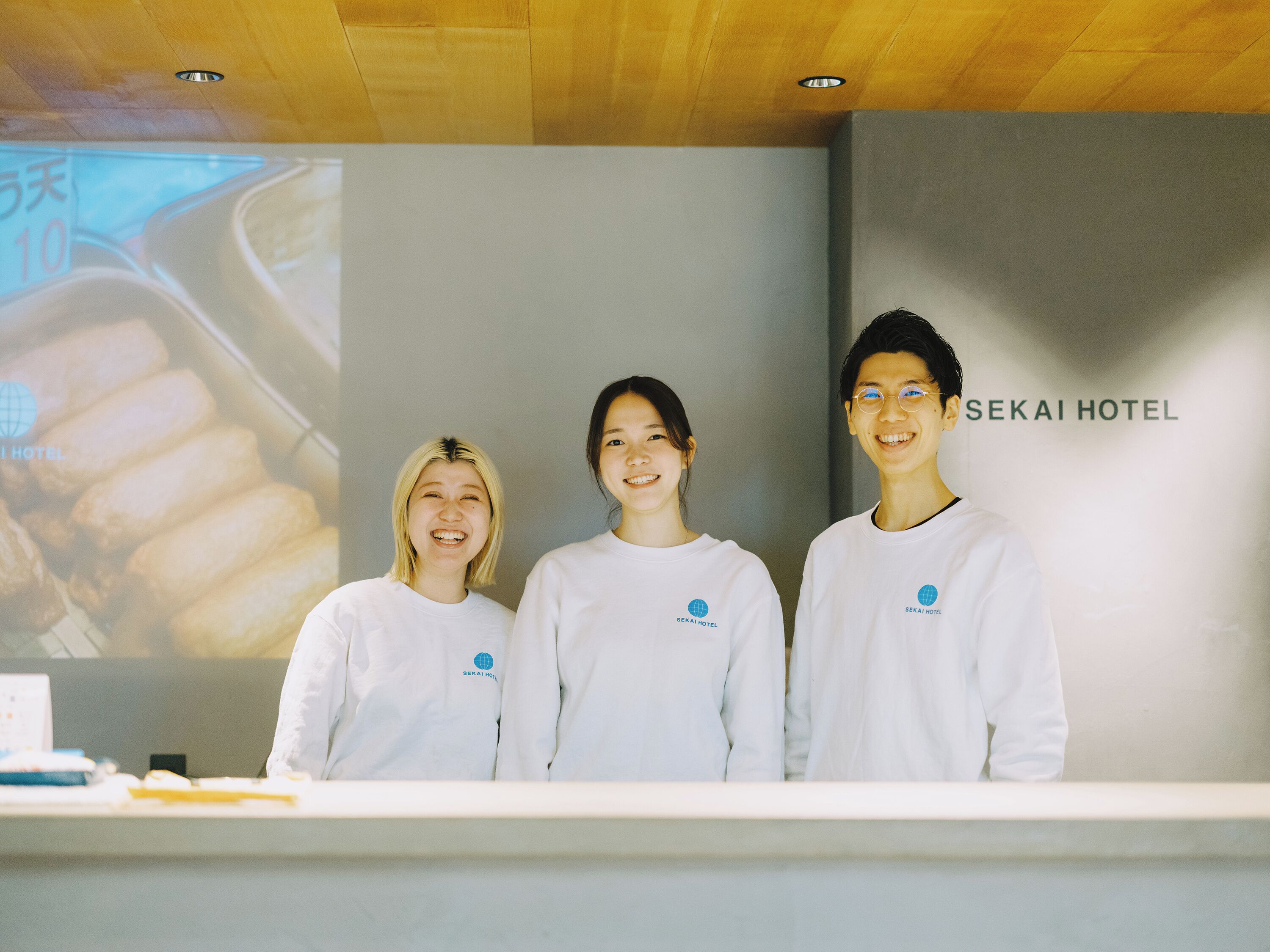 Three individuals standing in front of a reception desk with a large screen displaying a food image and the text 'SEKAI HOTEL'.