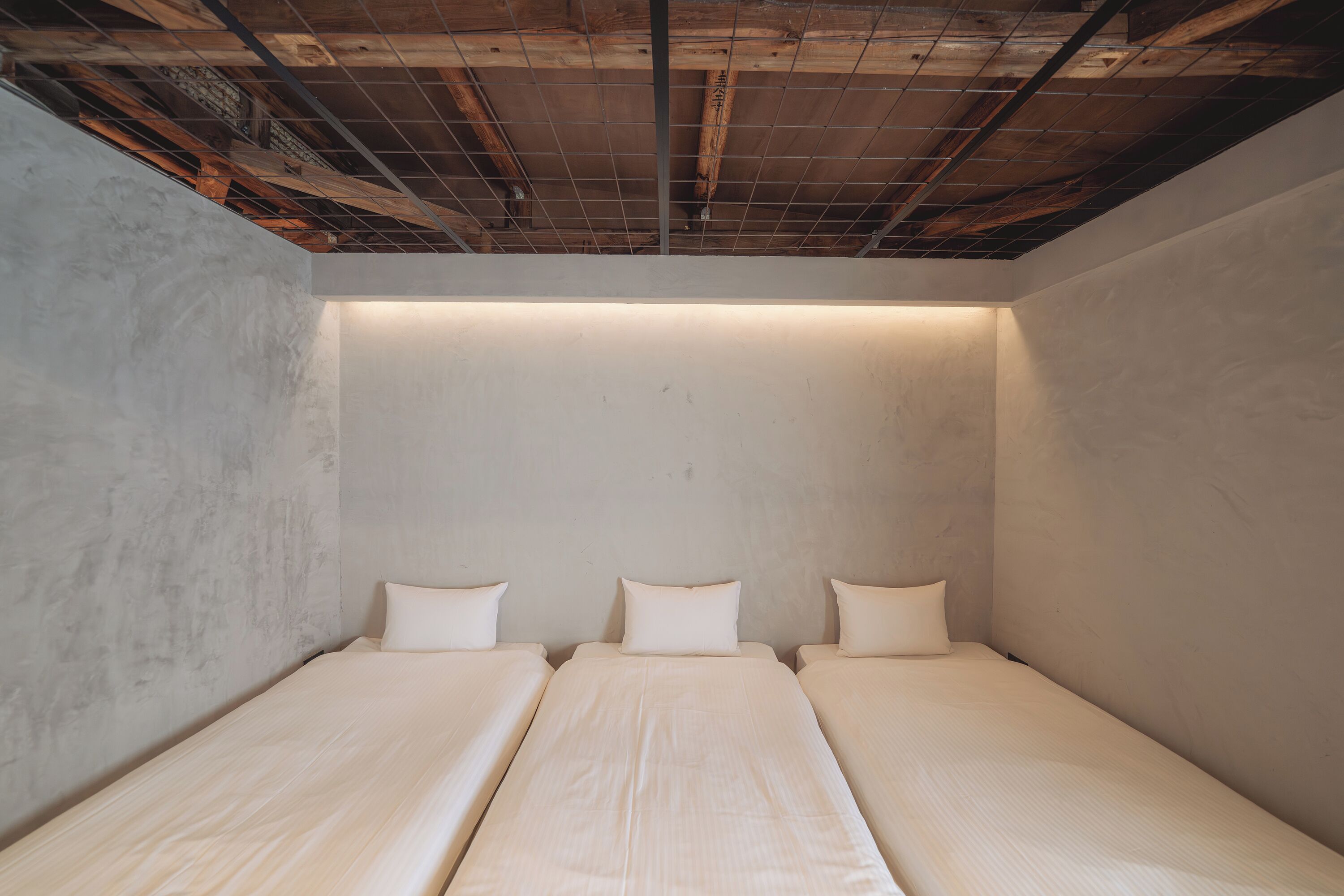 A room with three single beds aligned against a plain wall, with a visible wooden ceiling structure.