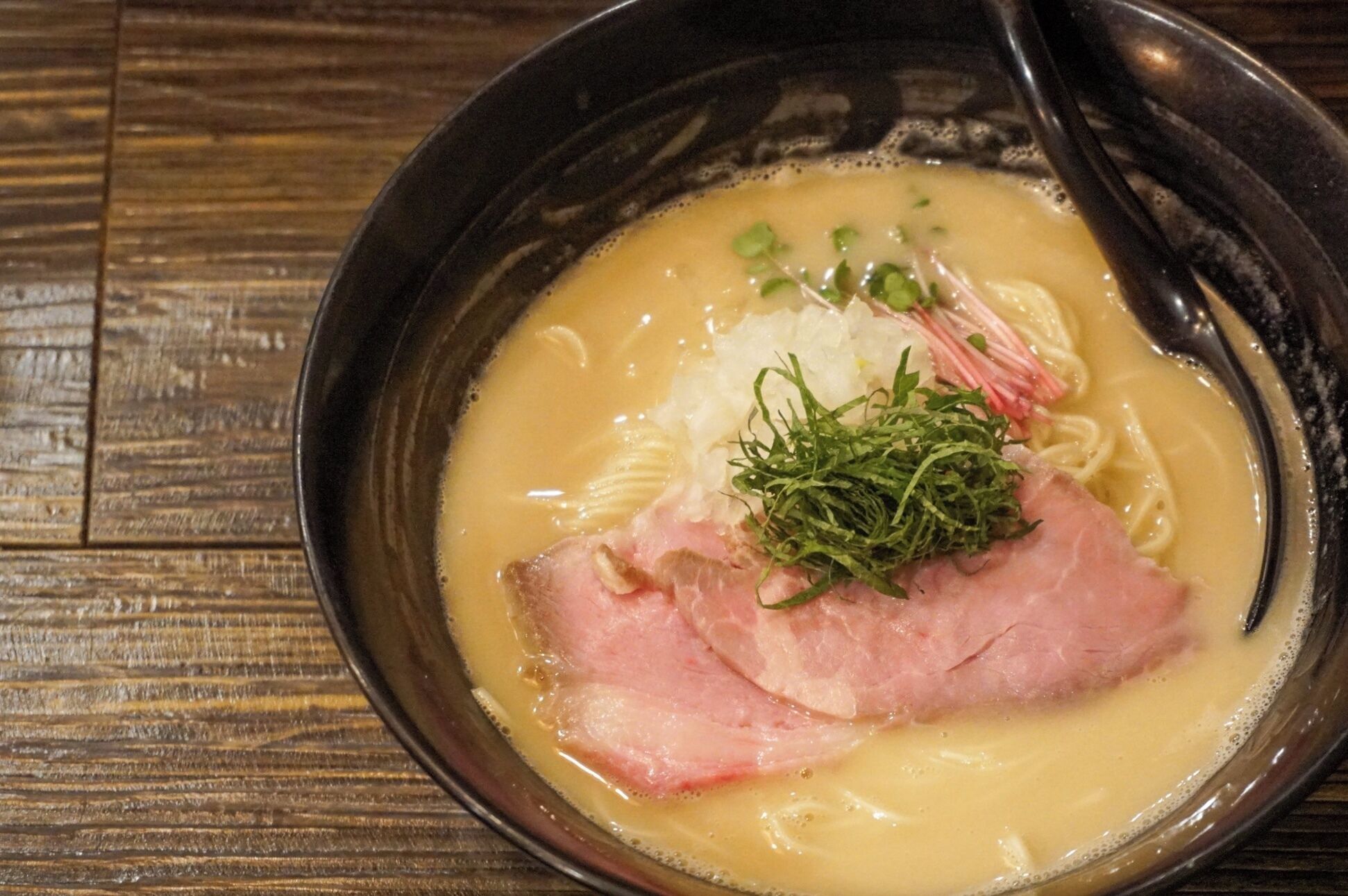 A bowl of ramen with noodles, pork, and toppings on a wooden table.
