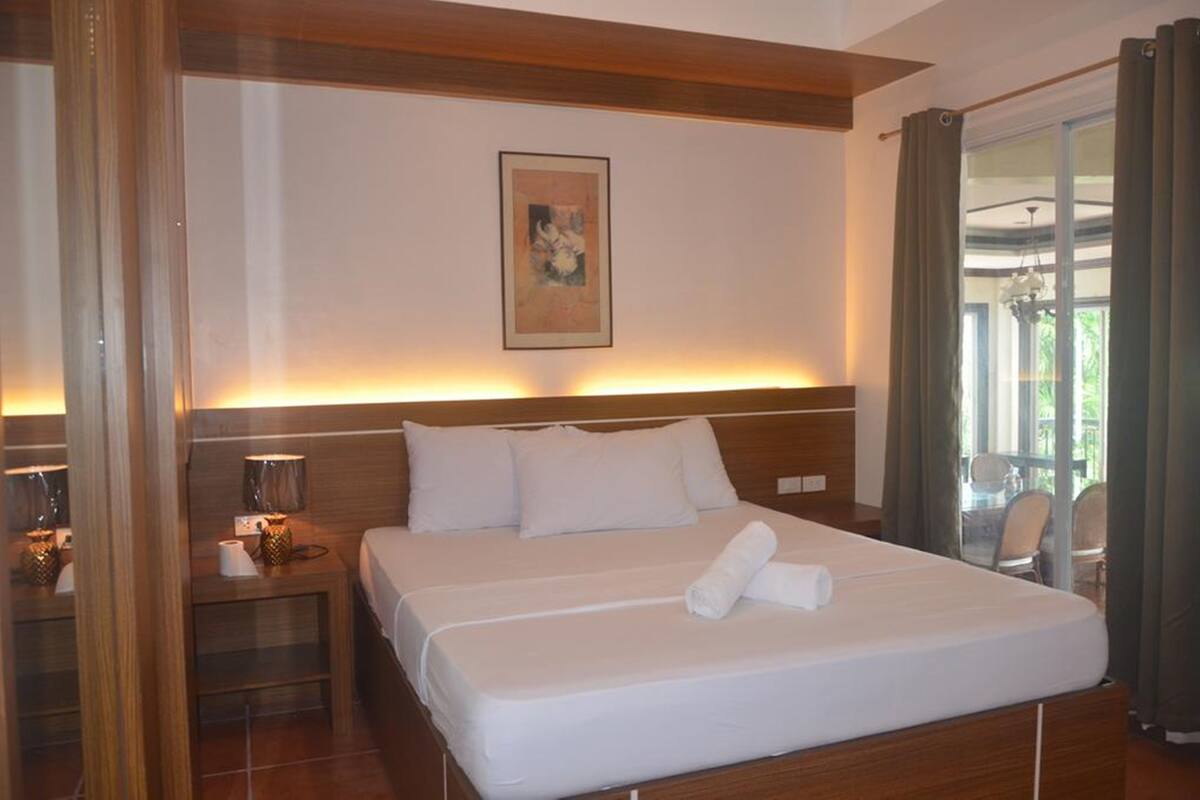 Une chambre d’hôtel avec un grand lit, une table de chevet avec une lampe, un tableau au mur et une vue sur l’extérieur par une fenêtre.