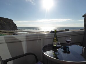 Un balcon avec une table, une bouteille de vin et deux verres. La vue comprend une plage et une falaise.