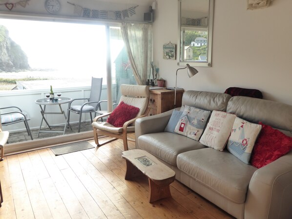 Un salon confortable avec un canapé, un fauteuil et une petite table. Il y a une fenêtre donnant sur la mer et un balcon avec des chaises.