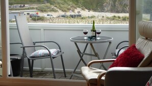 A balcony with a view of the beach, a table with two glasses of wine, and a chair with a red pillow. - Beach side Accommodation, Porthtowan, SATURDAY CHANGEOVER ONLY (Porthtowan)