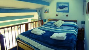 A bedroom with a bed, two bedside lamps, a window with curtains, and a painting on the wall. - Beach side Accommodation, Porthtowan, SATURDAY CHANGEOVER ONLY (Porthtowan)