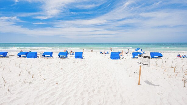 Una playa de arena blanca y cielo azul despejado. Hay sillas y sombrillas azules. Un letrero dice 'Bay Off the Dunes'.