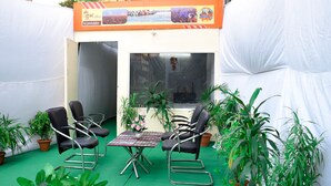 An outdoor seating area with two black chairs and a small table, surrounded by potted plants. - Kumbh Canvas (Prayagraj)