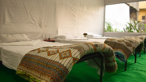 A room with a green carpet, white beds, and a window with a view of plants. - Kumbh Canvas (Prayagraj)