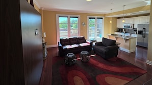 A spacious living room with a dark wood floor, a brown leather sofa, a grey armchair, and two black ottomans. There is a large window with a view of trees and a balcony. - 6th Floor Condo in The Dickson with View of Old Main (Fayetteville)