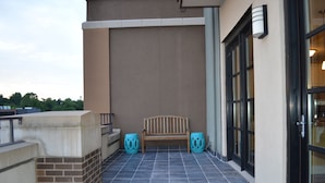 A balcony with a wooden bench and two decorative stools, a tiled floor, and a building facade with large windows and a door. - 6th Floor Condo in The Dickson with View of Old Main (Fayetteville)