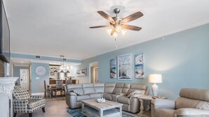 A spacious living room with a ceiling fan, a sectional sofa, a patterned armchair, a coffee table, and a dining area in the background. - Beautifully Condo BOOK TODAY (Navarre)