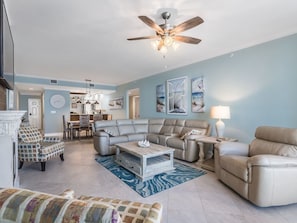A spacious living room with a ceiling fan, a sectional sofa, a patterned armchair, a coffee table, and a dining area in the background.