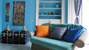 Ein Zimmer mit einem türkisfarbenen Sofa, einem Korbstuhl und einer blauen Wand. An der Wand hängen zwei Gemälde und ein blauer Vorhang.