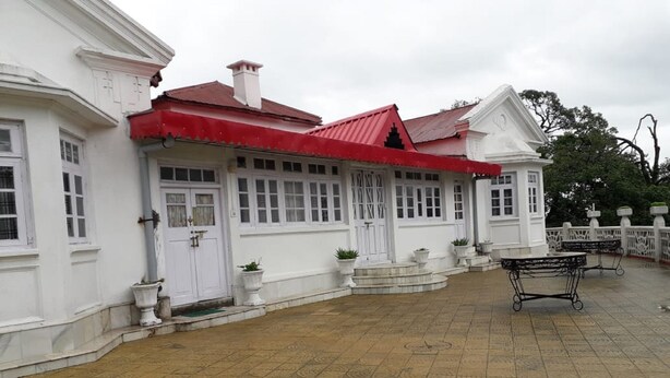 Un bâtiment blanc avec un toit rouge, une allée couverte et une petite construction à la façade blanche.