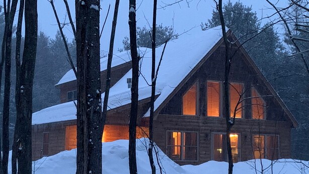 Une cabane en rondins dotée de nombreuses fenêtres et d’un toit recouvert de neige, entourée d’arbres.