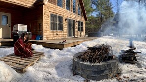 Une cabane en bois dotée de grandes fenêtres, d’une terrasse et d’un foyer extérieur, le tout au milieu de la neige.