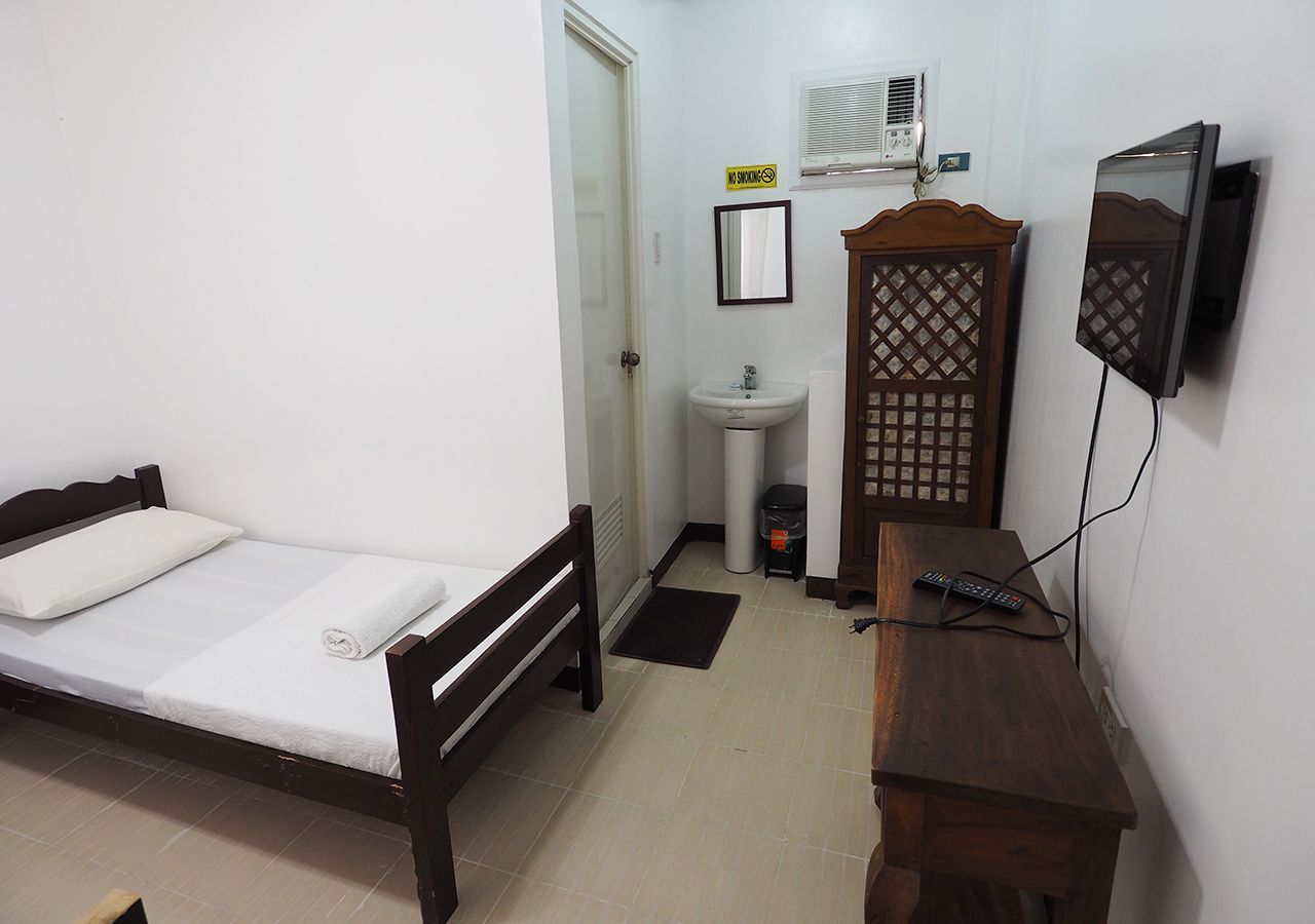 Una habitación pequeña y limpia con una cama individual, una mesita de noche, un armario de madera, un lavabo pequeño y un televisor montado en la pared.