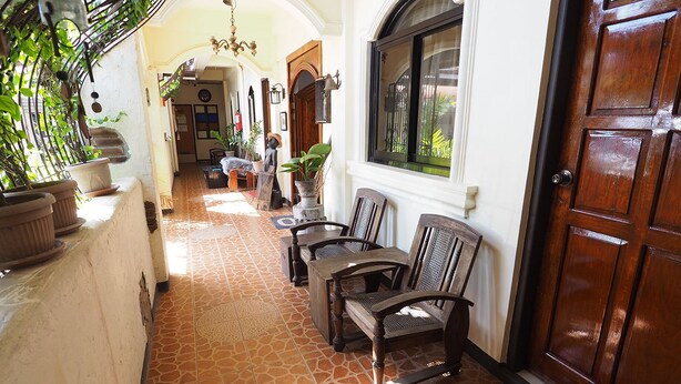 A corridor with a tiled floor, wooden chairs, a red mat, and a wooden door. - Balay Travel Lodge (Paoay)