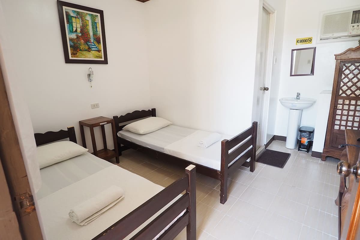 A room with two single beds, a small bedside table, a wall-mounted light, a painting, a sink, and a door with a wooden lattice design.