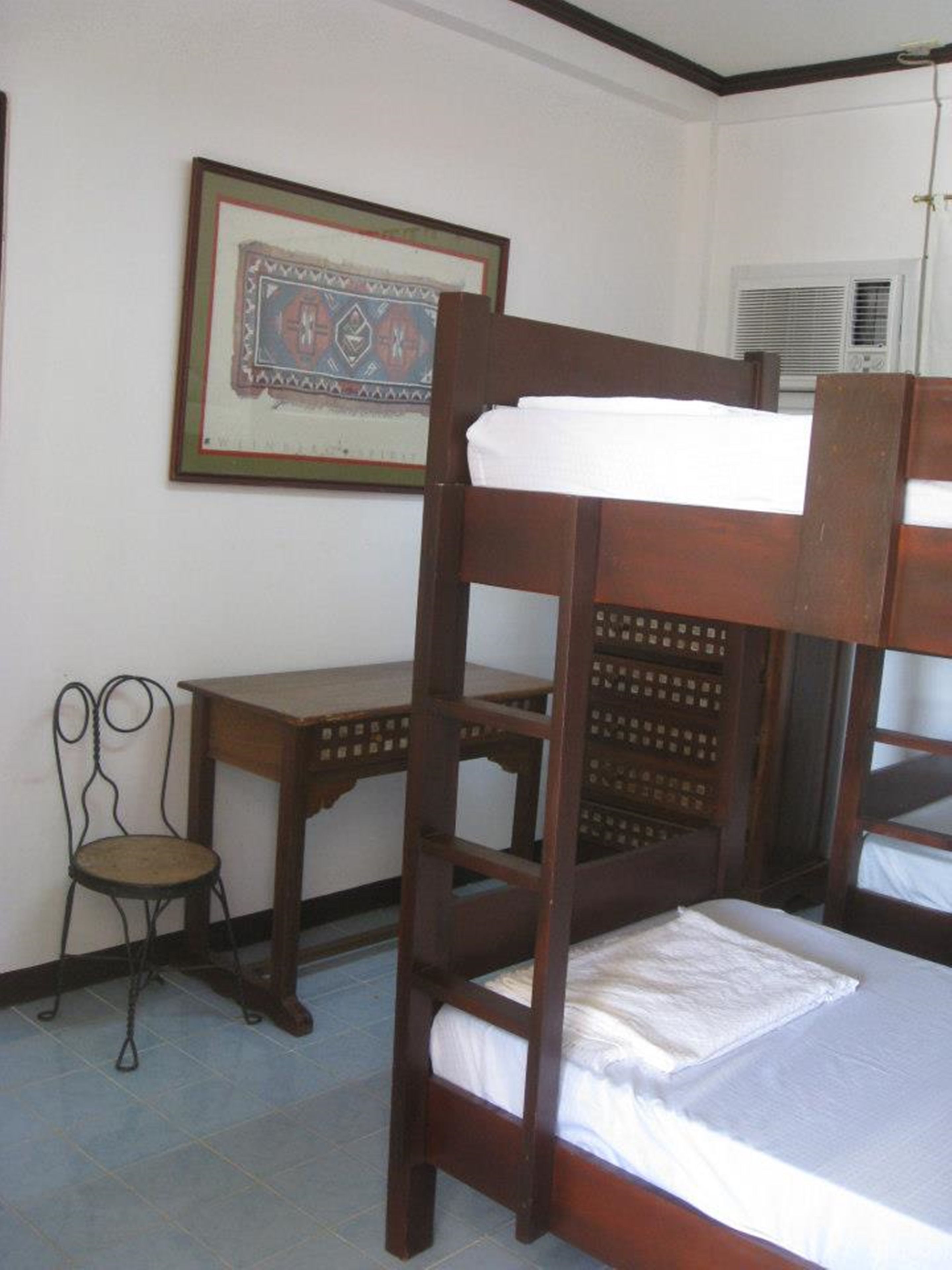 A bunk bed with a wooden frame and white bedding, a small wooden desk, a chair, and a framed picture on the wall.