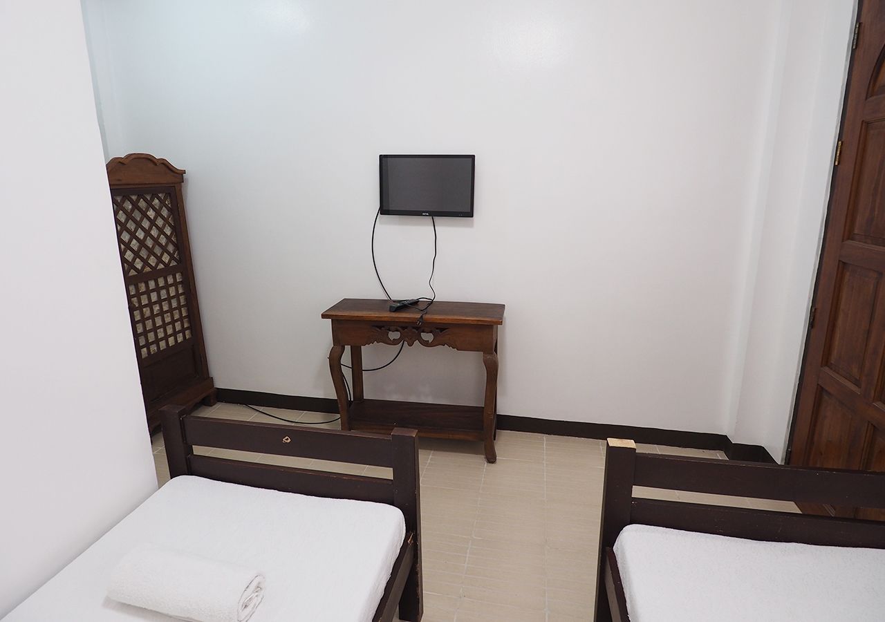Una habitación pequeña y sencilla con dos camas, una mesa de madera y un televisor colgado en la pared.