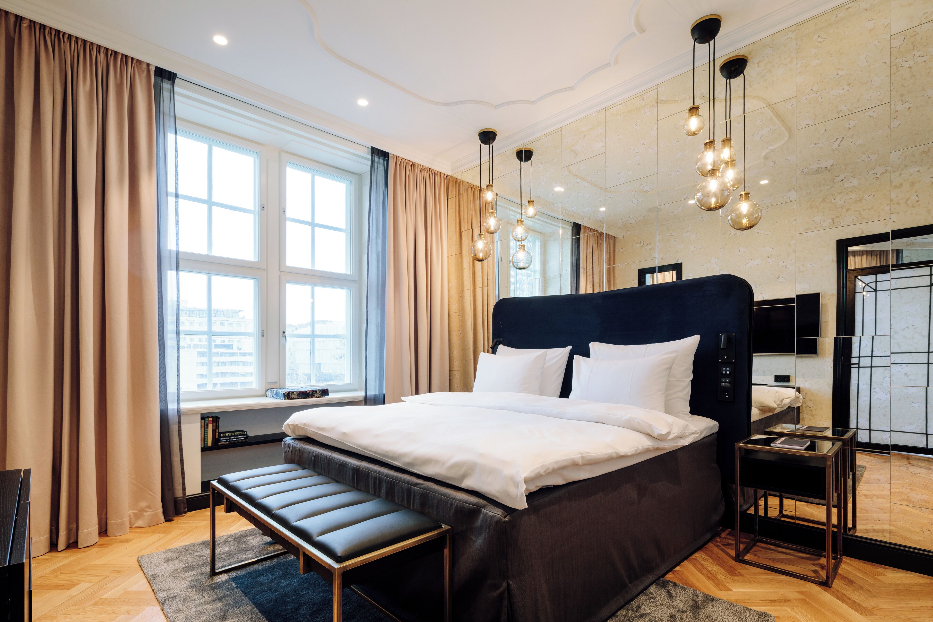 Una habitación de hotel moderna con una cama grande, una mesita de noche, un banco, un televisor y una ventana con cortinas.