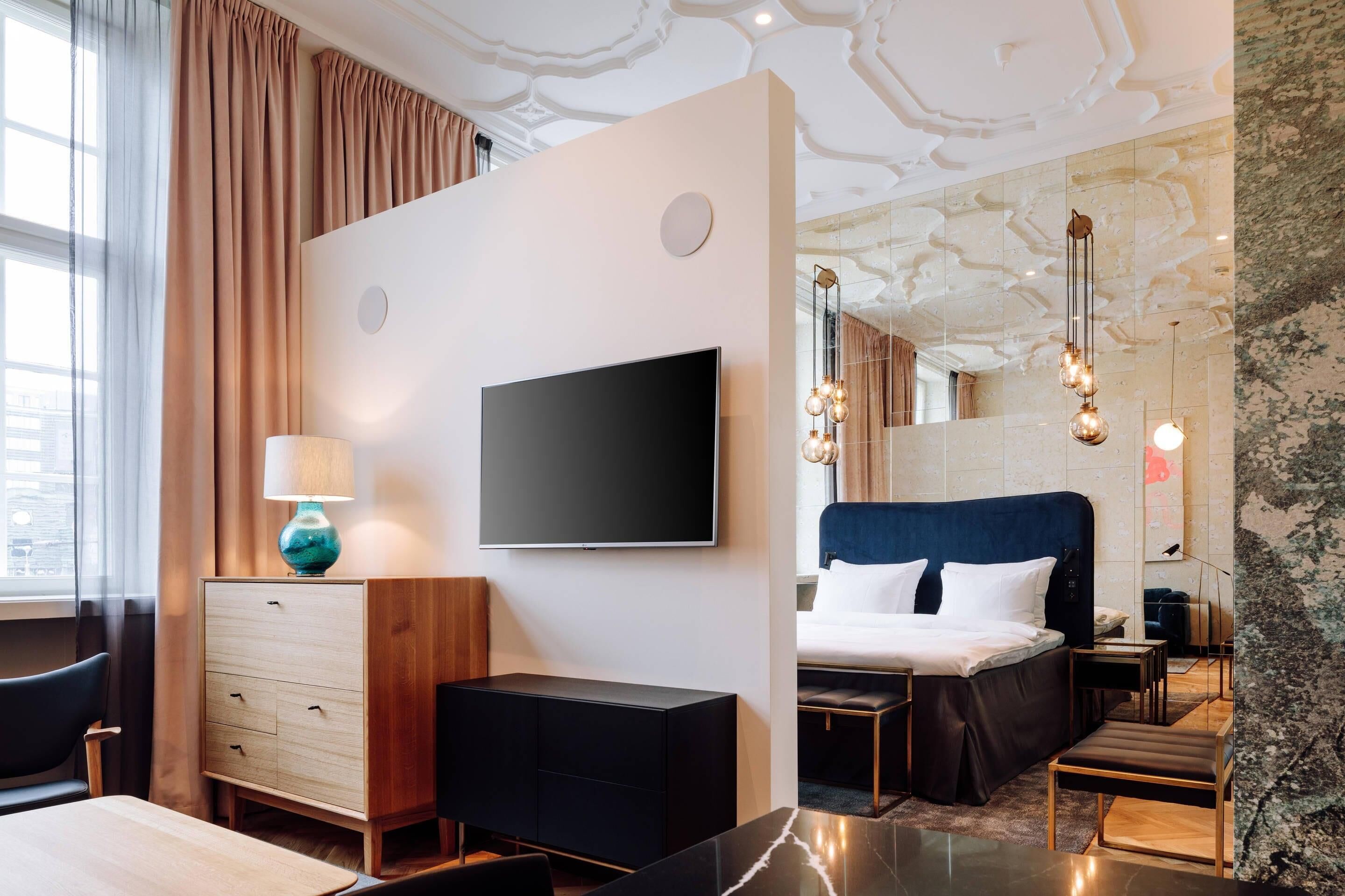 Quarto de hotel moderno com televisão de tela plana, um armário de madeira, uma escrivaninha, uma cama com cabeceira escura e um lustre.