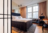 A modern hotel room with a large bed, a desk, and a view of the city through large windows.