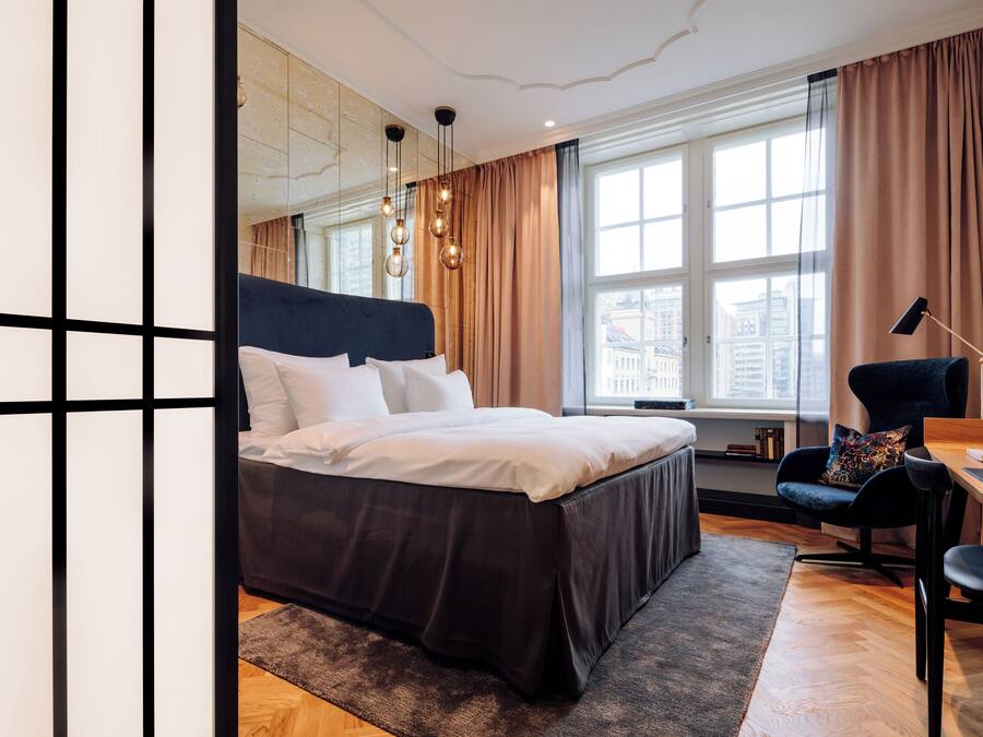 A modern hotel room with a large bed, a desk, and a view of the city through large windows.