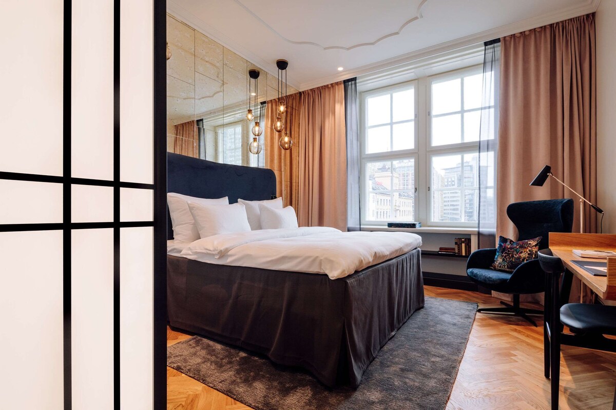 Una habitación de hotel moderna con una cama grande, un escritorio, una silla y vistas a la ciudad a través de amplios ventanales.