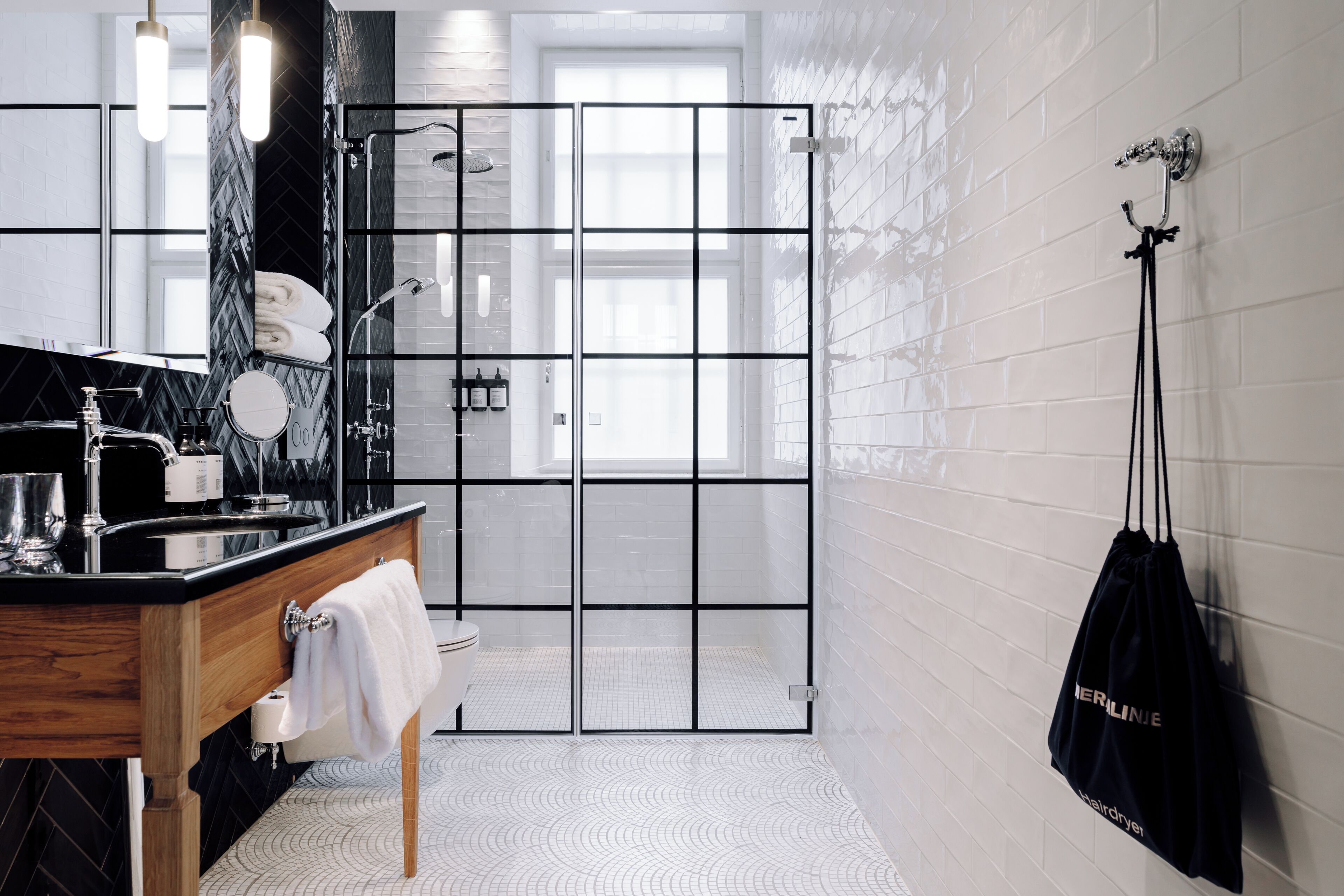 Un baño moderno con ducha de azulejos blancos y negros, tocador de madera y una bolsa colgada.