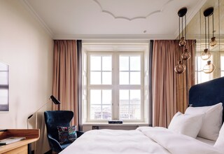 A hotel room with a large bed, a desk with a lamp, a chair, and a window with curtains.