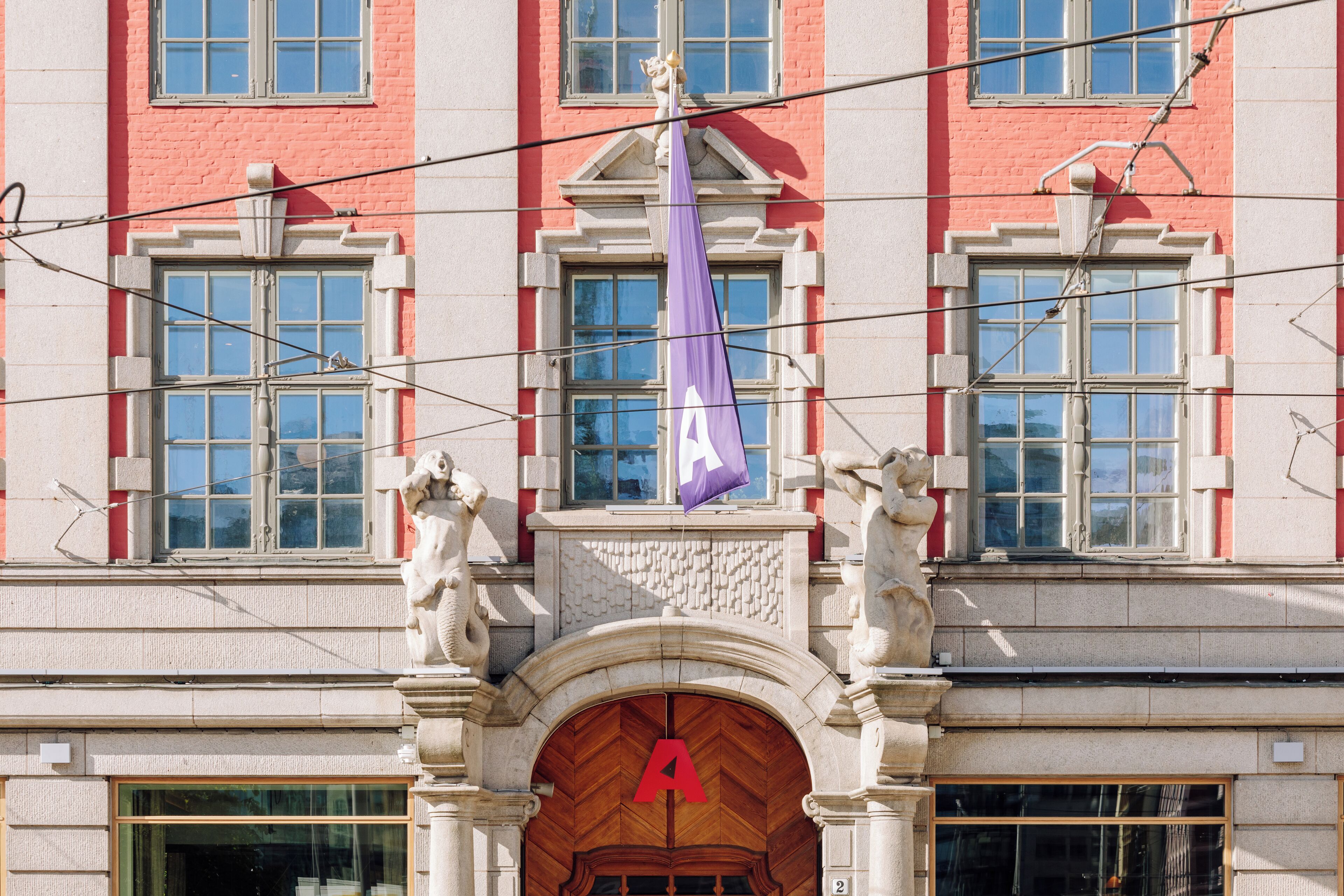 A building with a prominent red letter 'A' on the entrance, a purple flag, and classical architectural details including statues and a stone archway.