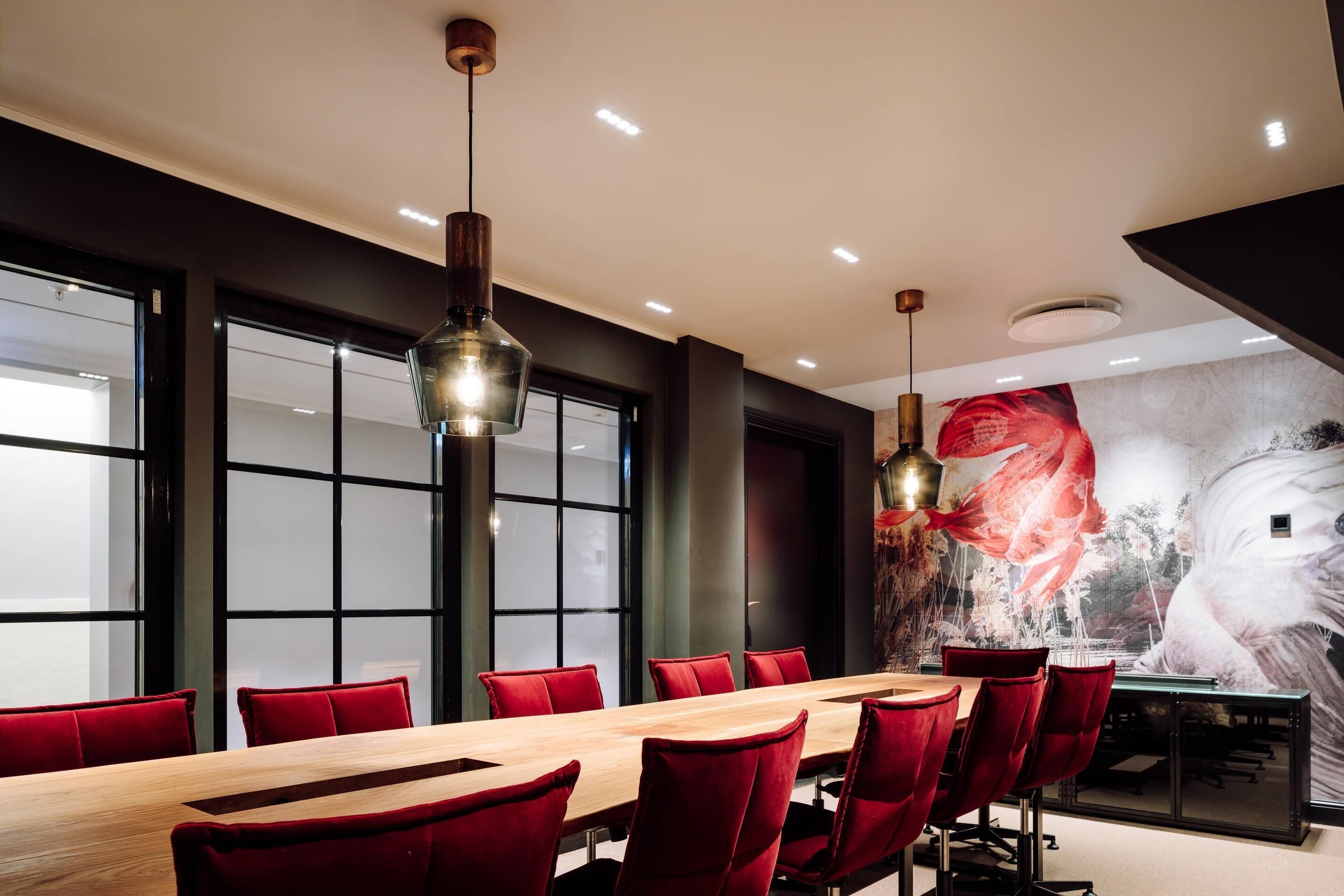 Une salle de conférence avec une longue table, des chaises rouges et des luminaires suspendus.