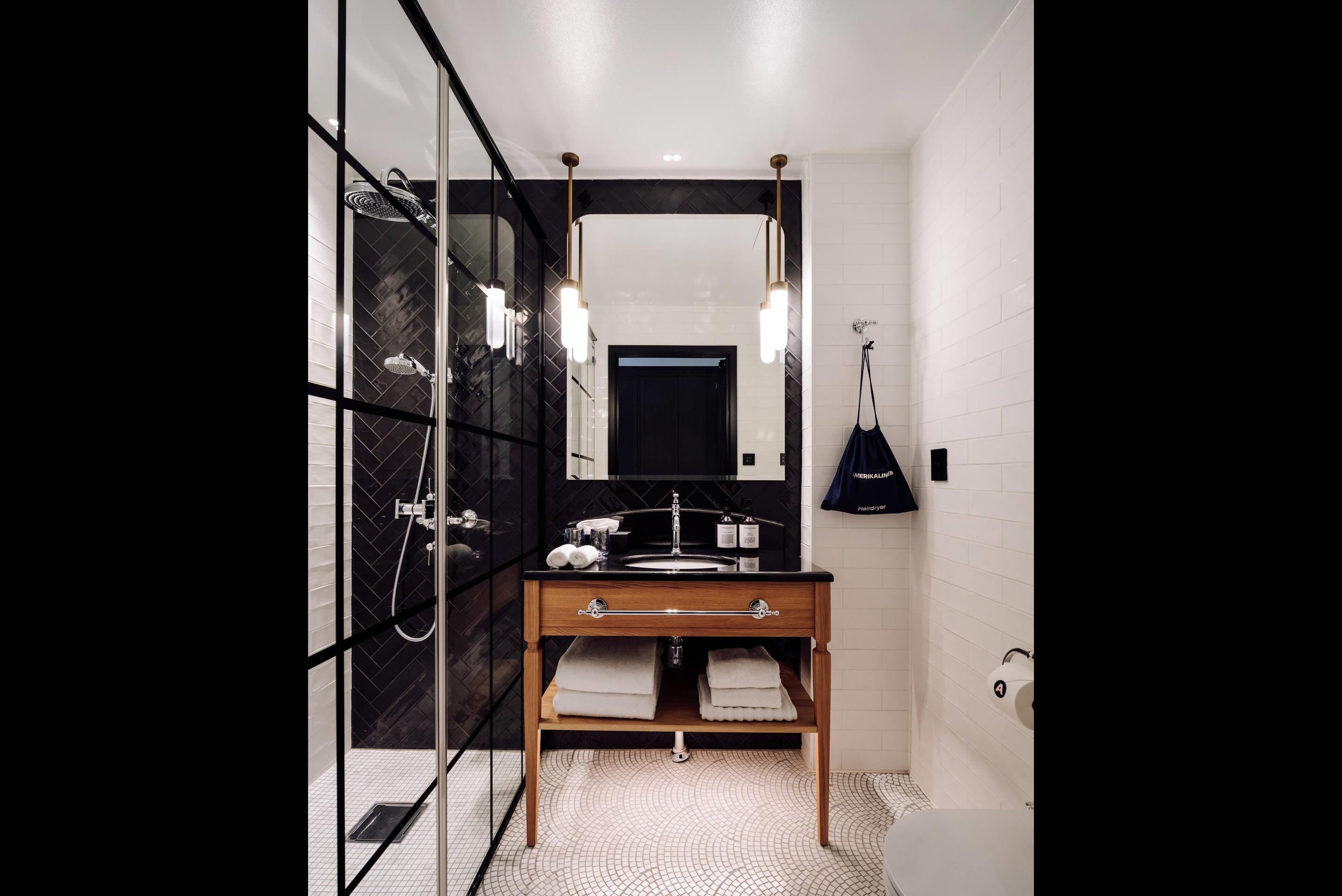 Un baño moderno con cabina de ducha de vidrio, tocador de madera con espejo y lámpara colgante negra.