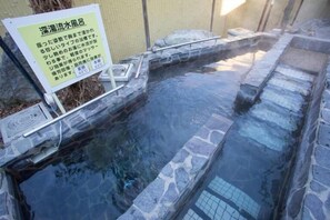 Une zone de sources chaudes avec des marches en pierre menant à l’eau, un panneau avec du texte en japonais et un mur de pierre en arrière-plan.