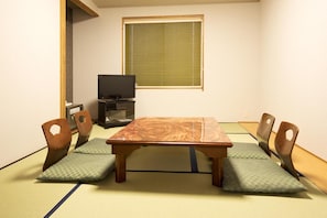 Une pièce de style japonais traditionnel, avec un sol recouvert de tatamis, une table basse, quatre chaises en bois, un téléviseur posé sur un meuble et une fenêtre équipée de stores.