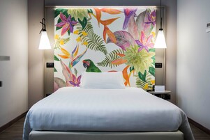 Camera d'albergo con un letto, due lampade sui comodini e un muro decorato con un murale floreale tropicale.