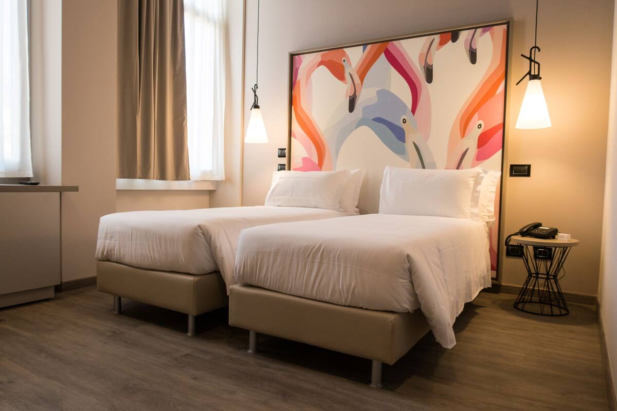 A hotel room with two beds, a small table with a phone, and a colorful abstract painting on the wall.