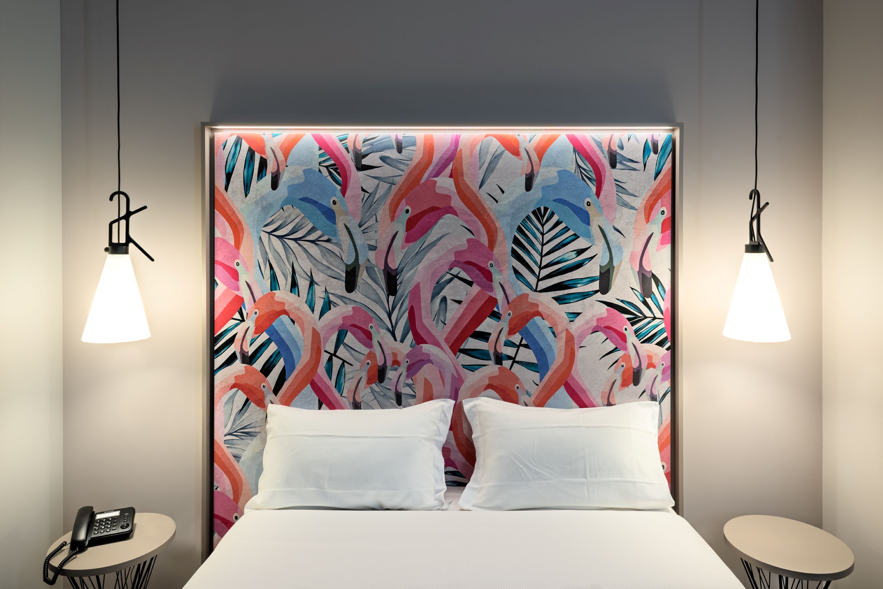 Une chambre d’hôtel avec un lit, des oreillers blancs, un téléphone sur une petite table et un mur orné d’un motif tropical aux couleurs vives.
