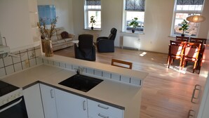 A modern kitchen with white cabinets, a black sink, and a built-in oven. There is a dining area with a table and chairs, and a living room with a sofa and a coffee table. - Abild Hotel (Tonder)