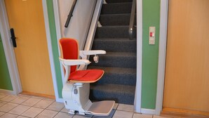 A wheelchair-accessible chair with a red seat and footrest, positioned near a staircase with green walls and beige flooring. - Abild Hotel (Tonder)