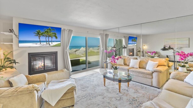 Un salon spacieux avec une cheminée, une table basse en verre et un grand canapé. Deux grandes fenêtres offrent une vue sur la plage et les palmiers.