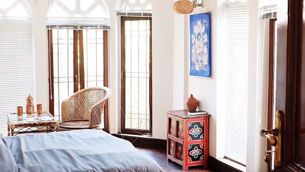 Una habitación con una cama, un sillón de mimbre, una mesita con objetos decorativos, una lámpara de pared y un mueble de almacenamiento colorido.
