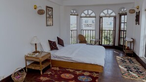 Un dormitorio con una cama grande, una mesita con lámpara, una silla de mimbre y vistas a través de ventanas de arco.